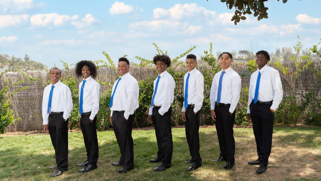 Seven young Black men dressed in white long sleeve button down shirts with blue ties, black slacks, and black dress shoes.